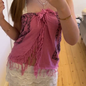 Rosa bandeau tubtopp med fransar - Säljer en rosa bandeau tubtopp med svarta mönster och fransar längs nederkanten. Toppen knyts framtill och har ett bohemiskt paisleymönster. Perfekt för sommardagar och festivaler. Materialet känns lätt och luftigt.