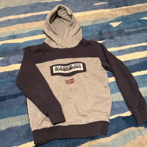 Napapijri hoodie i grått och mörkblått - Snygg hoodie från Napapijri i grått och mörkblått med broderad logga och norsk flagga på bröstet. Tröjan har huva, långa ärmar och ribbade muddar. Perfekt för dig som gillar streetwear och vill ha en stilren look.