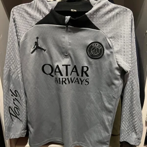 PSG långärmad träningströja Jordan - Snygg långärmad fotbollströja från PSG och Jordan i ljusgrått med svarta detaljer. Tröjan har halv dragkedja, Paris-logga på bröstet och diskret mönster i tyget. Tillverkad i lätt och ventilerande material, perfekt för träning eller match.