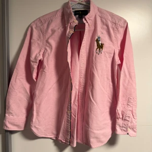 Rosa skjorta från Ralph Lauren M - Snygg rosa skjorta från Ralph Lauren med klassisk button-down krage och stor broderad polospelare på bröstet. Skjortan är långärmad, har knappar framtill och är tillverkad i mjuk bomull. Perfekt för dig som vill ha en stilren och preppy look.