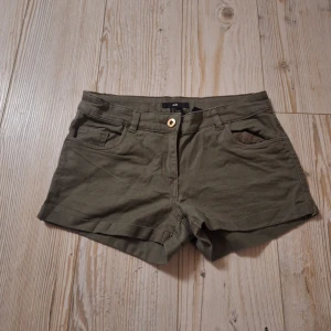 Olivgröna shorts med paljettdetaljer H&M - Snygga olivgröna shorts från H&M med coola rhinestones-detaljer på bakfickorna. Klassisk femficksmodell med knapp och dragkedja framtill. Perfekta för varma dagar och ger en extra touch med glittriga detaljer baktill.