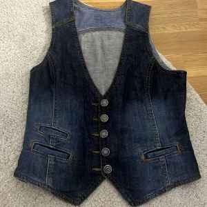 Mörkblå jeansväst med knappar - Snygg mörkblå jeansväst med fem silverfärgade knappar framtill och två fickor med dekorativa sömmar. Västen har v-ringad hals och ett justerbart bälte bak. Insidan har randigt foder längst ner. Perfekt för att ge din outfit en cool touch. Köpte på Zara för 399 och säljs för 299kr 