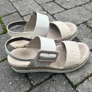 Beige sandaler med flätad detalj Rieker - Snygga sandaler från Rieker i beige med breda remmar, en flätad detalj framtill och stor spänne på sidan. Sulan har ett mönstrat kantband och är platt för extra komfort. Perfekta för varma dagar och enkel att matcha med sommarens outfits.