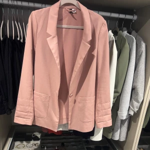 Ljusrosa kavaj med fickor - Snygg ljusrosa kavaj med klassisk krage och två stora fickor framtill. Enkelknäppt design och raka ärmar med diskreta sömmar vid mudden. 