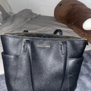 Svart Michael Kors handväska i skinn - Snygg svart handväska från Michael Kors i strukturerat skinn med guldfärgade detaljer och dragkedja. Väskan har två ytterfack, långa handtag och rymligt innerfack med flera småfickor. Perfekt för dig som vill ha plats för allt du behöver under dagen.