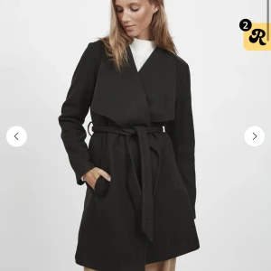 Svart omlott kappa med bälte - Snygg svart kappa med bred krage och omlott-design. Jackan har ett matchande bälte i midjan och två sidofickor. Långärmad och med stilren, minimalistisk look som passar perfekt till höst och vinter. Materialet ser ut att vara ull eller polyester.