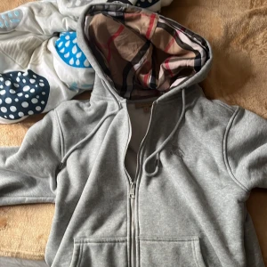Grå Burberry hoodie med rutig huva - Säljer en grå hoodie från Burberry med dragkedja och snörning. Insidan av huvan har det klassiska Burberry-rutiga mönstret i beige, svart och rött. Tröjan har broderad logga på bröstet och är tillverkad i mjuk bomull med fleecefoder. Perfekt för dig som gillar stilrena märkesplagg. Den passar S personer också 