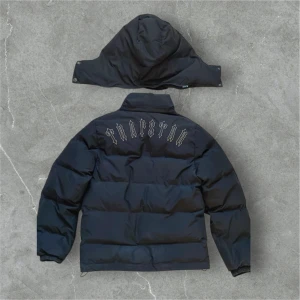 Svart pufferjacka från Trapstar XS - Svart pufferjacka från Trapstar med avtagbar huva och dragkedja framtill. Jackan har Trapstar-logga på bröstet och stor broderad text på ryggen. Puffig modell med elastiska muddar och två fickor med knappstängning. Perfekt för kalla dagar.