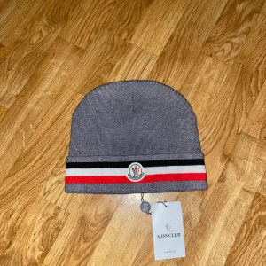 Moncler mössa  - Snygg grå Moncler mössa i ribbstickad bomull med klassiska Moncler-loggan framtill. Mössan har en bred kant med svarta, vita och röda ränder som ger en sportig vibe. Perfekt accessoar för att lyfta din vinterstil.