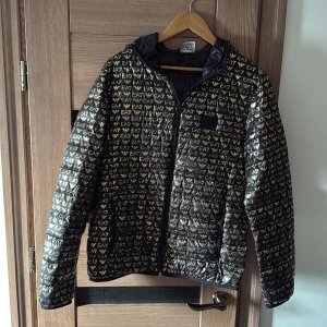 Svart och guld pufferjacka EA7 Armani - Snygg svart pufferjacka från EA7 Emporio Armani med guldiga logotyper över hela jackan. Jackan är helt oanvänd. 