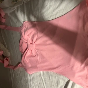 Rosa baddräkt med rosett - Supersöt ljusrosa baddräkt med breda axelband och en stor rosett framtill. Enkel och stilren design som ger en gullig vibe. Perfekt för stranden eller poolen i sommar!