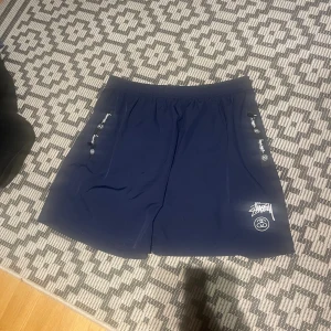 Mörkblå Stüssy shorts S/M - Snygga mörkblå shorts från Stüssy som passar S/M tillverkade i 100% polyester. De har elastisk midja, fickorna har stussy loggor på sig.