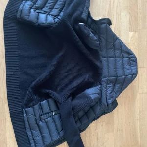 Svart Moncler pufferjacka med huva - Snygg svart pufferjacka från Moncler med stickade ärmar och quiltad kropp. Jackan har huva, dragkedja framtill och två fickor med knappstängning. Perfekt för dig som vill ha en stilren och varm jacka med exklusiv känsla.