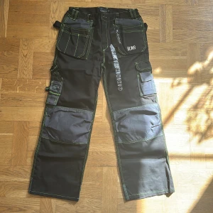 Svarta arbetsbyxor från Bluewear - Säljer ett par svarta arbetsbyxor från Bluewear med gröna kontrastsömmar och flera praktiska fickor. Byxorna har förstärkta partier på knän och ben, hällor för verktyg och BLWR-logga på fickan. Perfekta för dig som vill ha slitstarka och funktionella byxor.