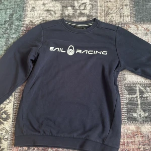 Mörkblå Sail Racing sweatshirt - Mörkblå sweatshirt från Sail Racing med vit logga framtill och liten logga bak i nacken. Klassisk rund halsringning med ribbade muddar och mjuk insida. Perfekt för dig som gillar stilren och sportig look.