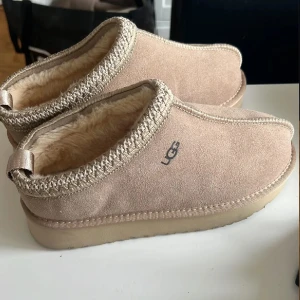 Beige UGG boots i mocka med foder - UGG boots i beige mocka med mjukt, fluffigt foder och grov sula. Skorna har rund tå, dekorativt band runt öppningen och UGG-logga på sidan. Perfekta för kalla dagar när du vill ha både stil och komfort. Aldrig använda 