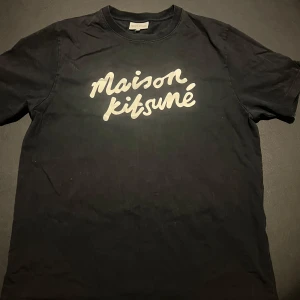 Maison Kitsuné svart t-shirt L - Svart t-shirt från Maison Kitsuné i storlek L med vit, stor logotyp på bröstet i coolt handskrivet typsnitt. Klassisk rund hals och korta ärmar. Tillverkad i mjuk bomull som är skön mot huden. Perfekt för dig som gillar stilrena och trendiga plagg.
