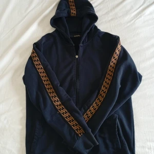 Mörkblå FJ Fendi hoodie med logoband - Snygg mörkblå hoodie från FJ Fendi med dragkedja och huva. Tröjan har bruna och svarta logoband längs ärmarna och över huvan, vilket ger en riktigt trendig look. Perfekt för dig som gillar streetstyle och vill sticka ut lite extra.