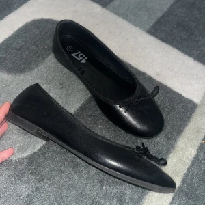 Svarta ballerinaskor med rosett - Klassiska svarta ballerinaskor från 157 med rund tå och liten rosett framtill. Skorna har en platt sula och är tillverkade i syntetmaterial med en lätt glansig finish. Perfekta för en stilren och enkel look. Använda 2 gånger