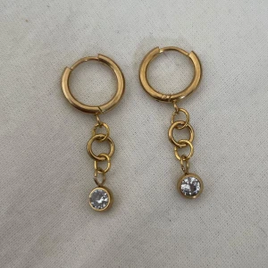 Guldiga örhängen med kedja och sten - Snygga guldiga örhängen med rund form och hängande kedjor. Längst ner sitter en glittrande klar sten som ger extra bling. Perfekt för dig som gillar stilrena detaljer med lite edge. Passar till alla tillfällen när du vill sticka ut. Rostfritt stål.