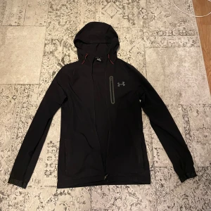 Svart vindjacka från Under Armour XS - Svart vindjacka från Under Armour med huva och röda dragsnören. Jackan har dragkedja framtill, en bröstficka med dragkedja och logga på bröstet. Ärmsluten har elastiska muddar med broderad text. Perfekt för blåsiga dagar och sportiga outfits. Ända felet med jackan ser ni på bild 5 o de är att jackan inte går att stänga eftersom de har gått av en del av dragkedjan där nere och fickan går inte att stänga  detta går o fixa hos nån smedjare för inte så dyrt. Annars är de ingenting fel på jackan 