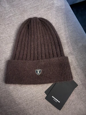Brun ribbad mössa Rockandblue - Snygg brun ribbstickad mössa från Rockandblue med uppvikt kant och metallmärke framtill. Modellen heter Felicia Neat och är tillverkad i mjukt stickat material som känns skönt mot huden. Perfekt för dig som vill ha en stilren och enkel look.