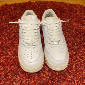 Nike Air Force 1 vita sneakers - Klassiska Nike Air Force 1 sneakers i helvitt skinn med perforerad tå, rund tå och platt sula. Snygg och tidlös modell med vita snören och diskret Nike-logga på sidan. Perfekta för dig som gillar en clean och stilren look.