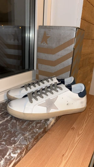 Golden Goose vita sneakers med stjärna - Golden Goose sneakers i vitt skinn med grå mockadetaljer och en ikonisk stjärna på sidan. Skorna har grå snörning, mörkblå häl och beige platt sula. Snyggt slitna detaljer och rund tå ger en cool och avslappnad vibe.