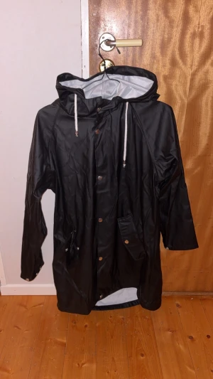 Svart regnjacka med huva och snörning - Svart regnjacka med huva och vita snören, perfekt för blöta dagar. Jackan har tryckknappar framtill, fickor med lock och en längre passform som skyddar mot regn. Materialet är syntet och jackan har en clean, enkel look.
