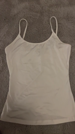 Vit linnetopp med smala axelband - Säljer en enkel och stilren vit linnetopp med smala axelband. Toppen har en rundad halsringning och ärmlös design, perfekt för varma dagar. Materialet känns mjukt och lätt mot huden. Passar dig som gillar minimalistisk och clean stil.