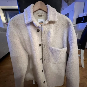Beige teddyjacka från Zara S-M - Mysig teddyjacka från Zara i beige med relaxed fit. Jackan har klassisk krage, knappstängning framtill och en stor bröstficka. Perfekt för dig som gillar chill och stilren look. Materialet är fluffigt och ger en skön vibe under höst och vinter.