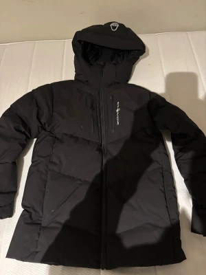 Svart pufferjacka Sail Racing, - Svart pufferjacka från Sail Racing med huva och flera dragkedjeförsedda fickor. Jackan har en stilren design med logga på bröstet och är perfekt för kalla dagar. Passformen är normal och materialet känns slitstarkt och varmt., använd ungefär 5 gånger köpt för 1 år sen ca, storlek 160