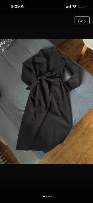 Svart lång kappa med bälte - Snygg svart kappa med bred krage och knytbälte i midjan. Lång modell med raka ärmar och stilren look, perfekt för dig som gillar minimalistisk stil. Materialet känns mjukt och har en matt finish. Passar dig som vill ha en klassisk vibe i garderoben.