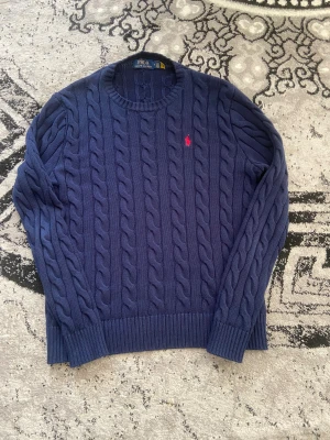 Marinblå stickad tröja Polo Ralph Lauren - Säljer en marinblå stickad tröja från Polo Ralph Lauren i storlek M. Tröjan har ett klassiskt kabelstickat mönster, rund hals och en röd broderad logga på bröstet. Perfekt för dig som gillar stilrena och tidlösa plagg.
