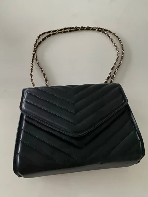 Svart quiltad axelväska med kedja - Snygg svart axelväska med quiltad design och guldfärgad kedja som axelrem. Väskan har ett lock med magnetlås och är tillverkad i skinnimitation. Perfekt storlek för det viktigaste och ger en cool vibe till din outfit.