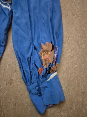 Blus - Säljer en blå blus från H&M i storlek 38 med färgglada blommor i rött, lila och beige. Blusen har långärmade ärmar med knappar vid mudden, v-ringad hals och dragkedja i ryggen. Materialet är mjukt och lätt.