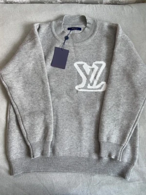 Grå stickad tröja Louis Vuitton - Säljer en grå stickad tröja från Louis Vuitton med stort vitt LV-logo framtill. Tröjan har ribbad krage, muddar och nederkant, samt långärmad passform. Materialet är mjukt och lyxigt, perfekt för dig som gillar designerplagg med clean look.