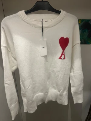 Vit stickad tröja från Ami - Vit stickad tröja från Ami med rund hals och ribbade muddar. På bröstet finns ett rött hjärta med bokstaven A, som är typiskt för märket. Tröjan har lång ärm och en clean, klassisk look som är lätt att matcha med jeans eller kjol.
