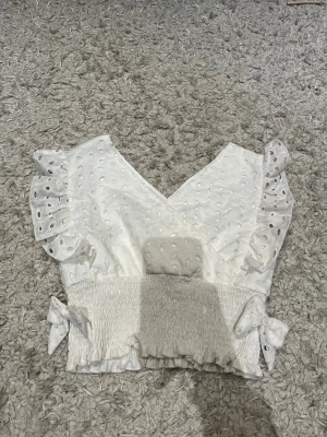 Vit blus med volang från SHEIN - Supersöt vit blus från SHEIN med broderade hålmönster, volangdetaljer på axlarna och v-ringad hals. Smockad midja med rosetter på sidorna och små volanger längst ner. Perfekt för dig som gillar romantisk och feminin stil.