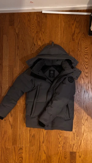 Grå pufferjacka från Canada Goose - Säljer en grå pufferjacka från Canada Goose med huva och snygga svarta detaljer. Jackan har dragkedja, tryckknappar och två stora fickor framtill. Märkets patch sitter på ärmen och insidan har svart foder. Perfekt för kalla dagar. S passar också på M.