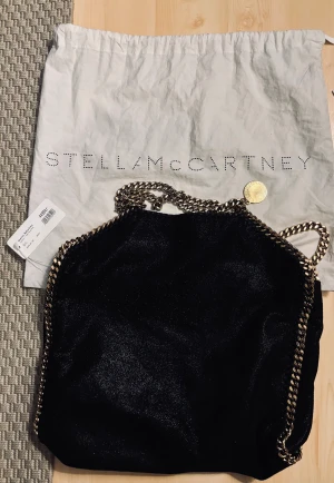 Svart axelväska Stella McCartney - Snygg svart axelväska från Stella McCartney med glansig finish och kraftiga guldfärgade kedjor längs kanterna och som axelrem. Väskan har en mjuk, slouchy form och en rund guldfärgad medaljong med logga. Perfekt för dig som gillar statement-accessoarer.