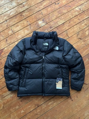 The North Face 1996 Retro Nuptse - Svart The North Face 1996 Retro Nuptse dunjacka i storlek M. Klassisk, varm och stilren vinterjacka med ikonisk boxig passform. Jackan är helt ny med prislapp kvar – perfekt för kalla vinterdagar