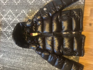 Svart Moncler pufferjacka med huva - Säljer en svart pufferjacka från Moncler med glansig finish och huva. Jackan har flera dragkedjeförsedda fickor, dubbla dragkedjor framtill och ikonisk Moncler-logga på ärmen. Insidan har ett coolt serietidningsmotiv. Perfekt för dig som gillar street och premium.