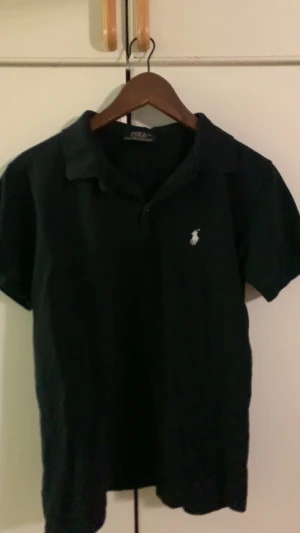Svart pikétröja från Polo Ralph Lauren - Svart pikétröja från Polo Ralph Lauren med klassisk krage och korta ärmar. Tröjan har den ikoniska vita broderade loggan på bröstet och är tillverkad i mjuk bomull som är skön mot huden. Perfekt för dig som gillar stilrena och tidlösa plagg.
