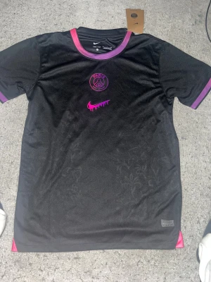 Svart PSG x Nike fotbollströja S - Säljer en svart PSG x Nike Dri-Fit fotbollströja i storlek S. Tröjan har korta ärmar, lila och rosa detaljer vid krage och ärmslut, samt PSG-logga och Nike swoosh i rosa framtill. Materialet är lätt och andas, perfekt för träning eller match.