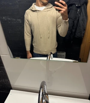 Beige stickad hoodie med snörning - Snygg beige stickad hoodie med huva och långa ärmar. Tröjan har snörning vid halsen och ribbade muddar vid ärmslut och nederkant. Perfekt för dig som gillar en chill och stilren look. Passar dig som vill ha en normal passform.