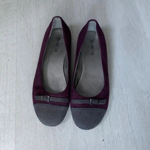 Vinröda ballerinaskor från Bonita - Snygga ballerinaskor i vinröd mocka från Bonita med mörkgrå tå och dekorativt band med liten spänne framtill. Skorna har platt sula och rund tå, vilket gör dem både stilrena och bekväma. Perfekta för dig som vill ha en klassisk men ändå trendig look.