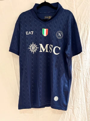 Napoli matchtröja EA7 marinblå - Snygg SSC Napoli matchtröja i marinblått med subtilt randigt mönster och tryckta loggor i vitt och guld. Tröjan har korta ärmar, V-ringad krage och är tillverkad i ventilerande polyester. MSC och klubbmärken på bröstet ger en autentisk look.