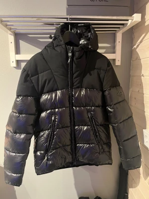 Svart pufferjacka från Replay - Snygg svart pufferjacka från Replay med glansiga paneler och huva. Jackan har två dragkedjeförsedda fickor fram, Replay-logga på ärmen och quiltad insida. Perfekt för kalla dagar med sin tjocka och fluffiga känsla. Säljer tyvärr denna då jag vuxit ur den