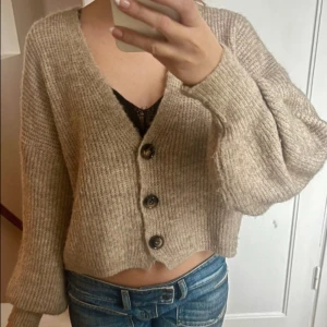 Beige stickad kofta med knappar - Säljer en beige stickad kofta med tre stora knappar framtill. Koftan är croppad och har långärmade, lite puffiga ärmar